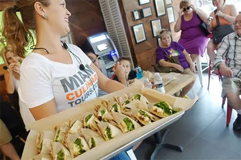 A woman is holding a tray of tacos in front of a group of people on a culinary tour.