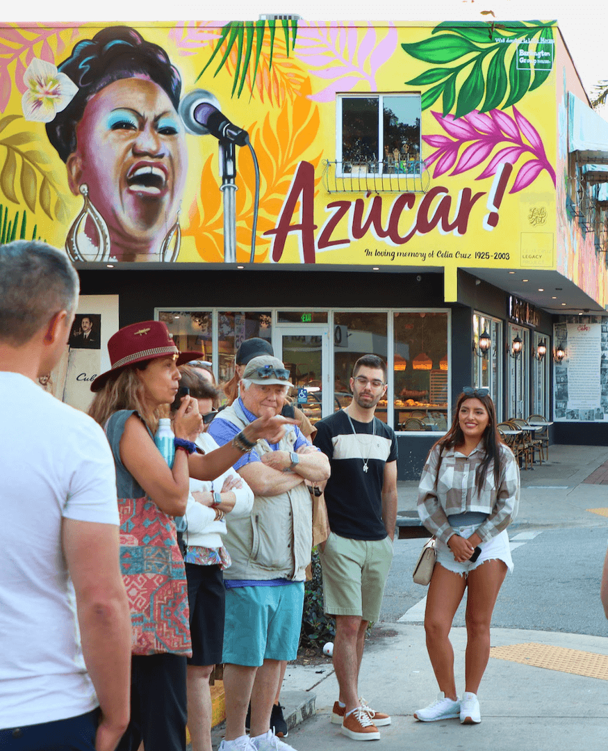 People gathered on a street with a colorful mural in the background commemorating cuban culture.