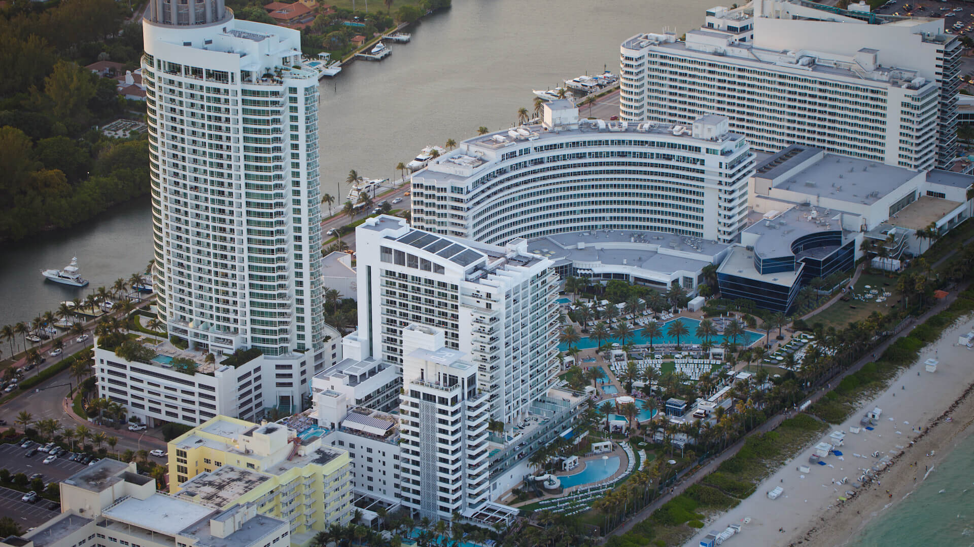 Aerial view of a large beachfront hotel complex with multiple white buildings, outdoor pools, palm trees, and the ocean on one side and a river on the other.