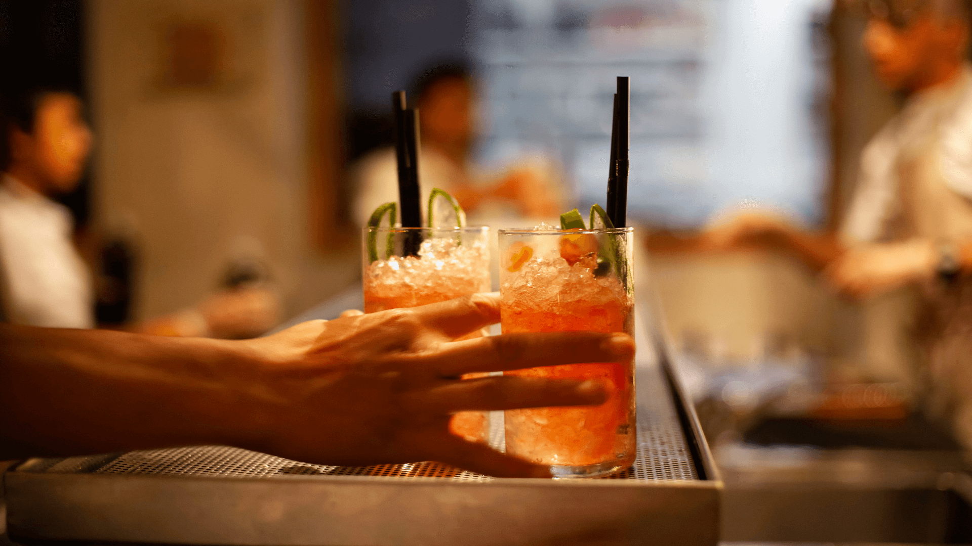 A person places two iced cocktails with lime slices and straws on a bar counter, with blurred people in the background.