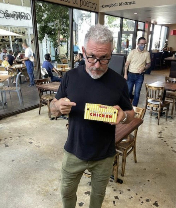 A man with gray hair and glasses stands in a Miami Design District restaurant holding a yellow striped box labeled "CHICKEN!" and points at it.