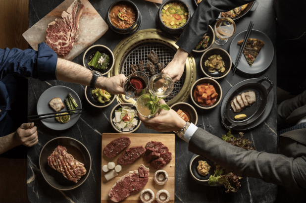 A table set with various Korean BBQ dishes and sides in the Miami Design District, with three people toasting drinks over a central grill—perfect inspiration for your next Dining Guide adventure.