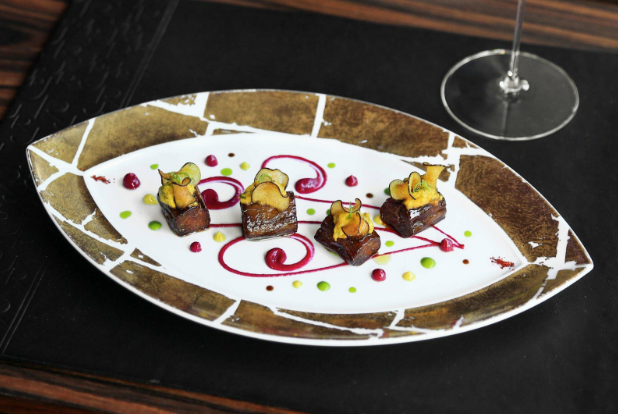 A gourmet dish of four small meat pieces topped with vegetables, artfully plated with colorful sauce dots and swirls on a decorative oval plate, offers a taste of the Miami Design District’s vibrant dining guide experience next to a wine glass.
