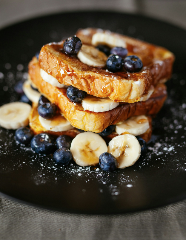 Three slices of French toast stacked on a black plate, topped with banana slices, blueberries, powdered sugar, and syrup—a must-try indulgence in the Miami Design District dining guide.
