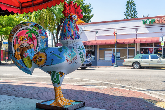 A large, colorful rooster statue with various painted designs stands on a sidewalk in Little Havana, Miami, in front of shops and parked cars on a sunny day.