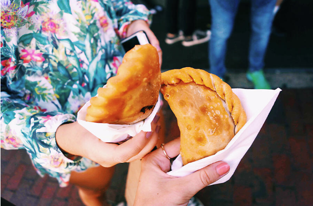 Two people holding fried empanadas in white napkins, one wearing a floral-patterned shirt, standing on a brick surface in Little Havana—perfect for any Miami cultural guide.