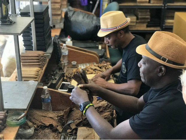 Two men wearing hats hand-roll cigars at a worktable in Little Havana, Miami, surrounded by tobacco leaves and cigar-making tools—a cultural guide to traditional craftsmanship.