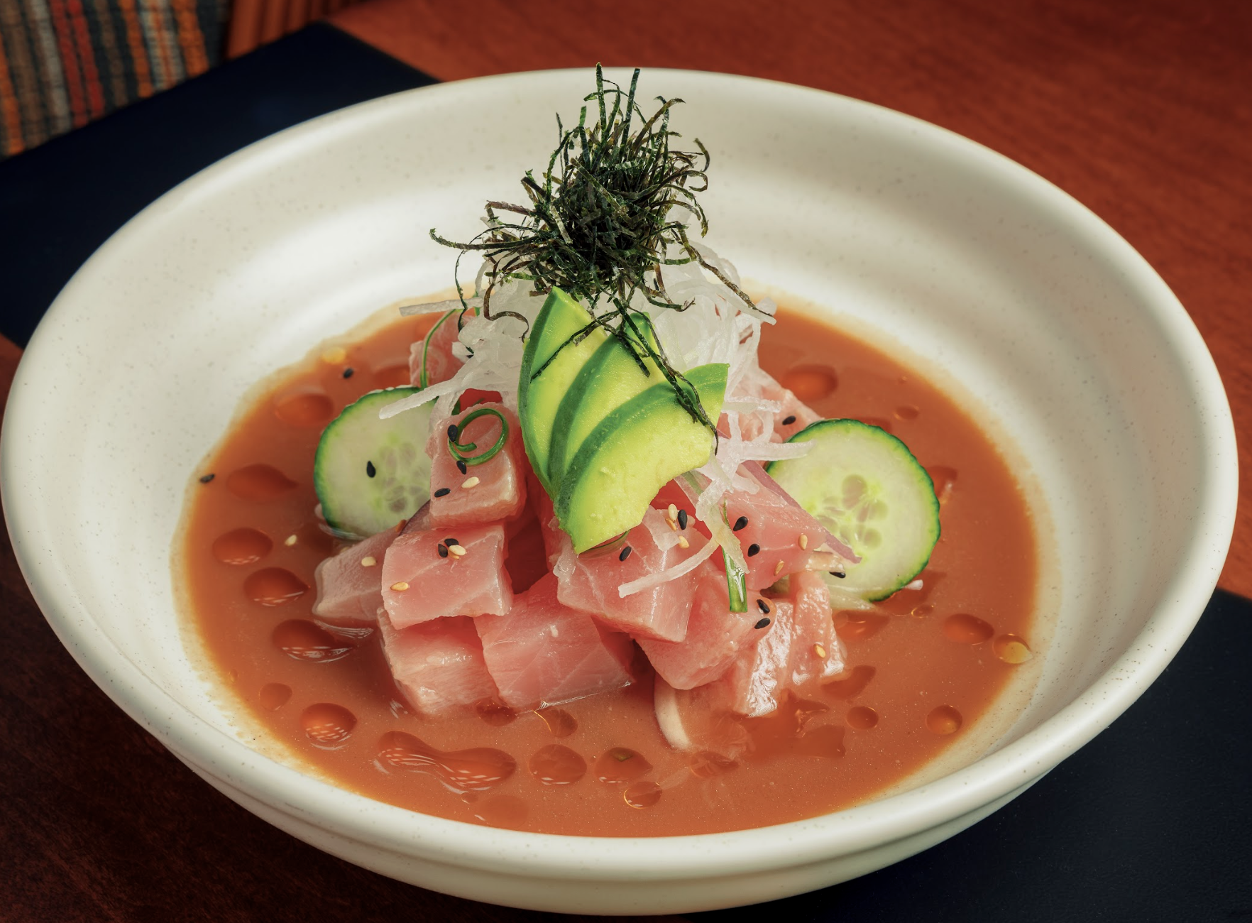 A white bowl with cubes of raw fish in a reddish sauce, topped with sliced cucumber, avocado, radish, and shredded seaweed—a fresh bite perfect for Miami June events and trendy food and drink happenings.